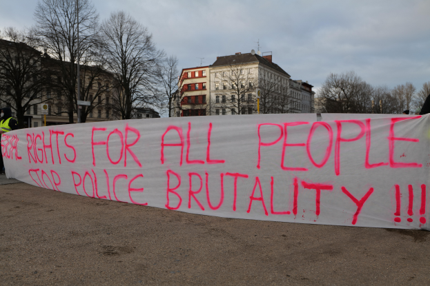 Eine Gruppe von Menschen hält ein Transparent mit der Aufschrift 'Rechte für alle Menschen Stoppt Polizeigewalt' auf dem Boden mit einer Straßenlaterne, einem Schild, Bäumen, Gebäuden mit Fenstern und einem bewölkten Himmel im Hintergrund.
