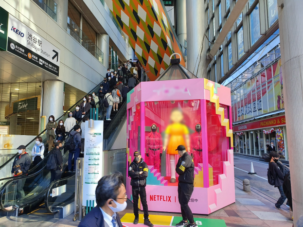 Eine Gruppe von Menschen steht vor einem Gebäude mit Glaswänden, Säulen und Texttafeln, einige tragen Masken, während eine Rolltreppe mit Menschen daneben läuft; ein pinker Kasten in der Mitte zeigt Schaufensterpuppen und trägt den Text "Netflix's new Netflix-Booth in Tokyo."