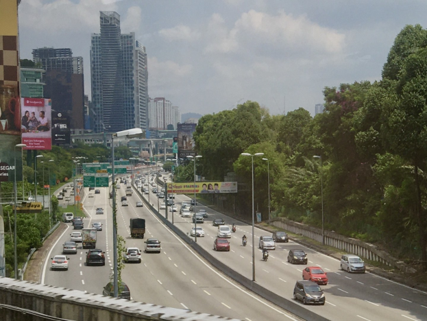 Eine vielbefahrene Autobahn mit hohen Gebäuden, Straßenlaternen, Leuchtreklamen, Bäumen und einem bewölkten Himmel.