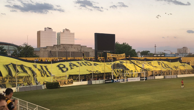 Ein Fußballfeld mit einer Gruppe von Menschen darauf, umgeben von einem Metallzaun, einem Torpfosten, einer Fahne mit der Aufschrift "Boca Juniors vs Real Sociedad", einigen Pflanzen, einem Anzeigebildschirm, einigen Pfosten, Baumrinde, einigen Bäumen, einigen Gebäuden und einem bewölkten Himmel.