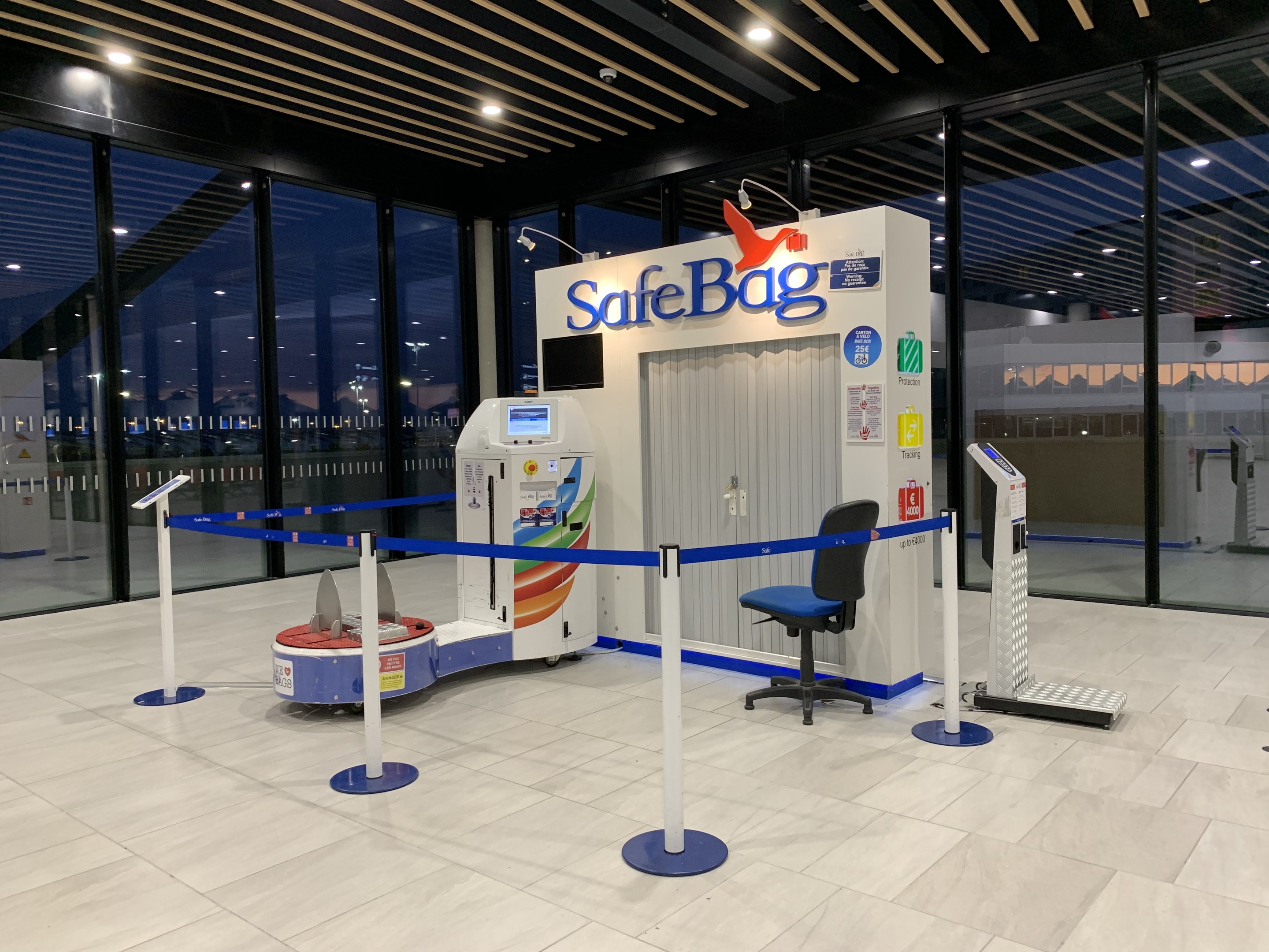 Sicherheitskontrolle für Gepäck am Flughafen mit einem Stuhl, Absperrgittern, Maschinen, einem Bildschirm, einer bedruckten Tafel, Glaswänden, Deckenleuchten und sichtbarem Boden.