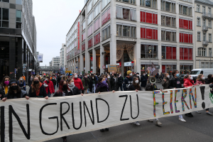 Eine Gruppe von Menschen mit Masken und einem Banner mit der Aufschrift "In Grund zu Feiern" steht vor Gebäuden mit Fenstern, Laternenmasten und Fahrzeugen, was auf eine Demonstration in Berlin, Deutschland, hindeutet.