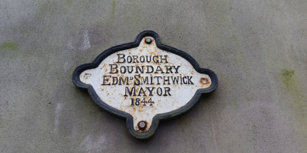 Metallschild an einer Gebäudeseite mit schwarzem Hintergrund und weißer fetter Schrift, die "Borough Boundary Edm Smithwick Mayor" lautet.