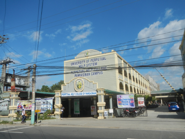 Die Universität Perpetual Help System in Pasig, mit Gebäuden, Fahrzeugen auf der Straße, Fußgängern auf dem Gehweg, Werbetafeln, Strommasten, Kabeln, Bäumen und einer bewölkten Himmel.