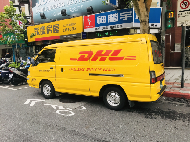 Gelbes DHL-Lieferfahrzeug auf der Straßenseite mit Gebäuden, Bäumen, Schildern, Mästen, Ampeln und einer Person auf dem Gehweg.