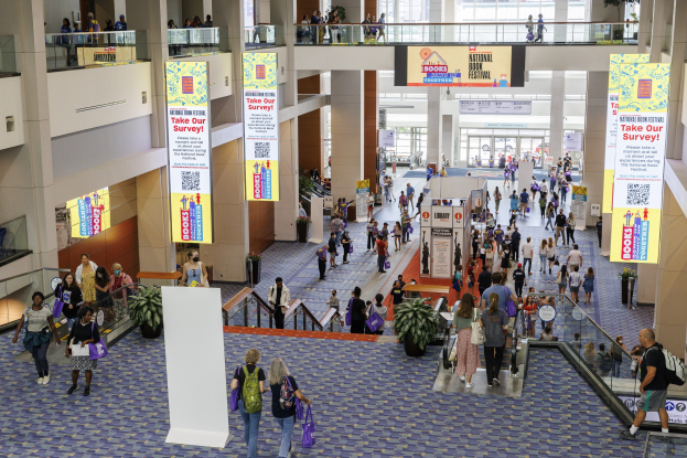 Eine große Gruppe von Menschen, die durch ein großes Gebäude mit Bannern, Topfpflanzen, Rolltreppen, Geländern, Säulen, bedruckten Schildern, Glastüren und anderen Gegenständen geht, einige tragen Taschen, wahrscheinlich nehmen sie am Nationalen Buchfestival teil.