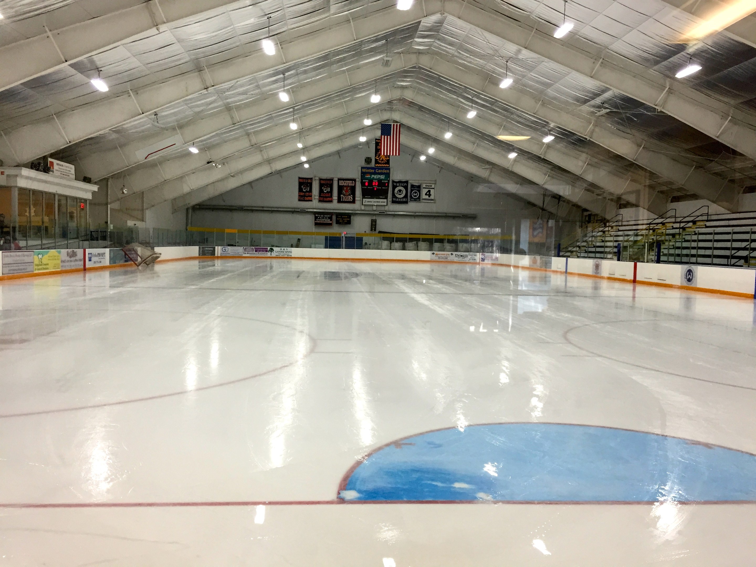 Großes Hallen-Eisstadion mit Deckenbeleuchtung, Werbetafeln, Treppen mit Geländern, einer Wand im Hintergrund und einigen Menschen, was auf eine Sportanlage hinweist, die likely für Hockey genutzt wird.