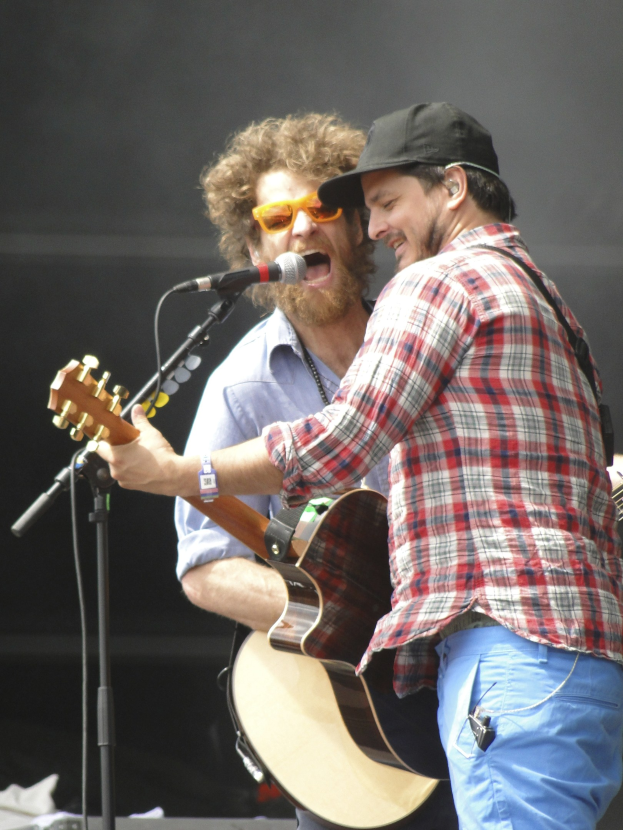 Zwei Männer mit Gitarren auf der Bühne, einer trägt ein rot-blau kariertes Hemd mit blauen Hosen, der andere eine Sonnenbrille und eine schwarze Mütze.