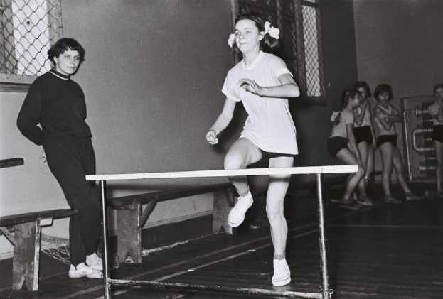 Schwarz-weißes Foto einer Frau in der Luft beim Überwinden eines Hindernisses in einer Halle mit Zuschauern und Bänken im Hintergrund.