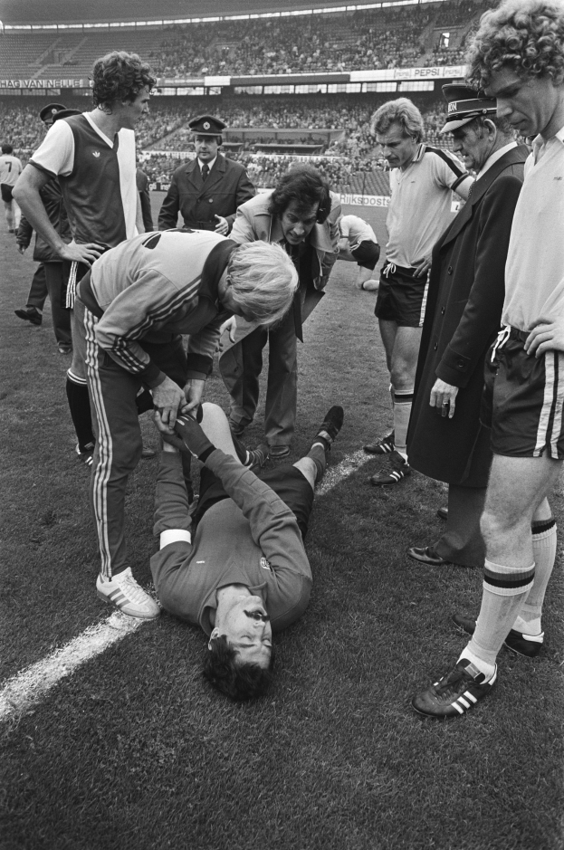 Schwarzes Bild eines Fußballspielers, der auf dem Boden liegt und von einer Gruppe Männer umgeben ist, mit einer Stadionkulisse im Hintergrund; der Spieler scheint verletzt zu sein.
