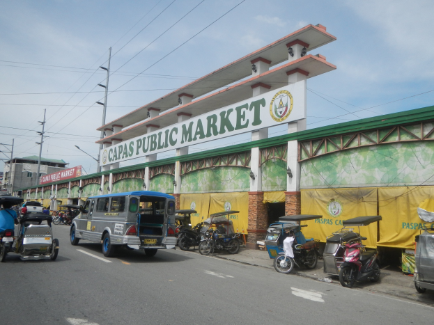 Lebendige Stadtstraße mit Autos, Motorrädern und Rikschas, die an dem Gebäude "Capas Public Market" vorbeifahren, Strommasten, Laternenmasten, Gebäude und bewölkter Himmel im Hintergrund.