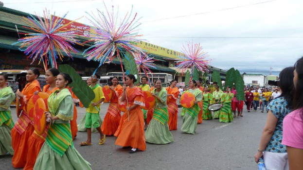 Eine Gruppe von Menschen in bunten Kostümen marschiert auf einer Straße während eines Karnevalsumzugs, mit einer Frau auf der rechten Seite, die eine Flasche hält, und Fahrzeugen, Gebäuden, Bäumen, Pfählen, Drähten und dem Himmel im Hintergrund.