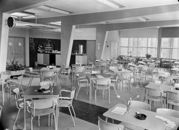 Schwarz-weiß-Foto einer Cafeteria mit Tischen und Stühlen, Tellern, Besteck, Gläsern und Blumenvasen auf den Tischen sowie einer Wand mit Türen, Fenstern und Deckenleuchten im Hintergrund.