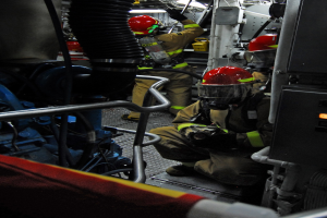 Feuerwehrleute in Helmen und Schutzausrüstung arbeiten an einem Löschfahrzeug mit Rohren und Gegenständen im Hintergrund, unter einem Licht an der Spitze.