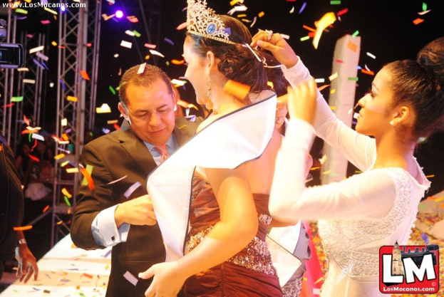 Ein Mädchen auf der Bühne bei einer Miss World Show, das eine Krone trägt, während ein anderer Mädchen dahinter die Krone anpasst und ein Mann in einem schwarzen Anzug vor ihr die Hand schüttelt, vor einem dunklen Hintergrund mit Metallrahmen.
