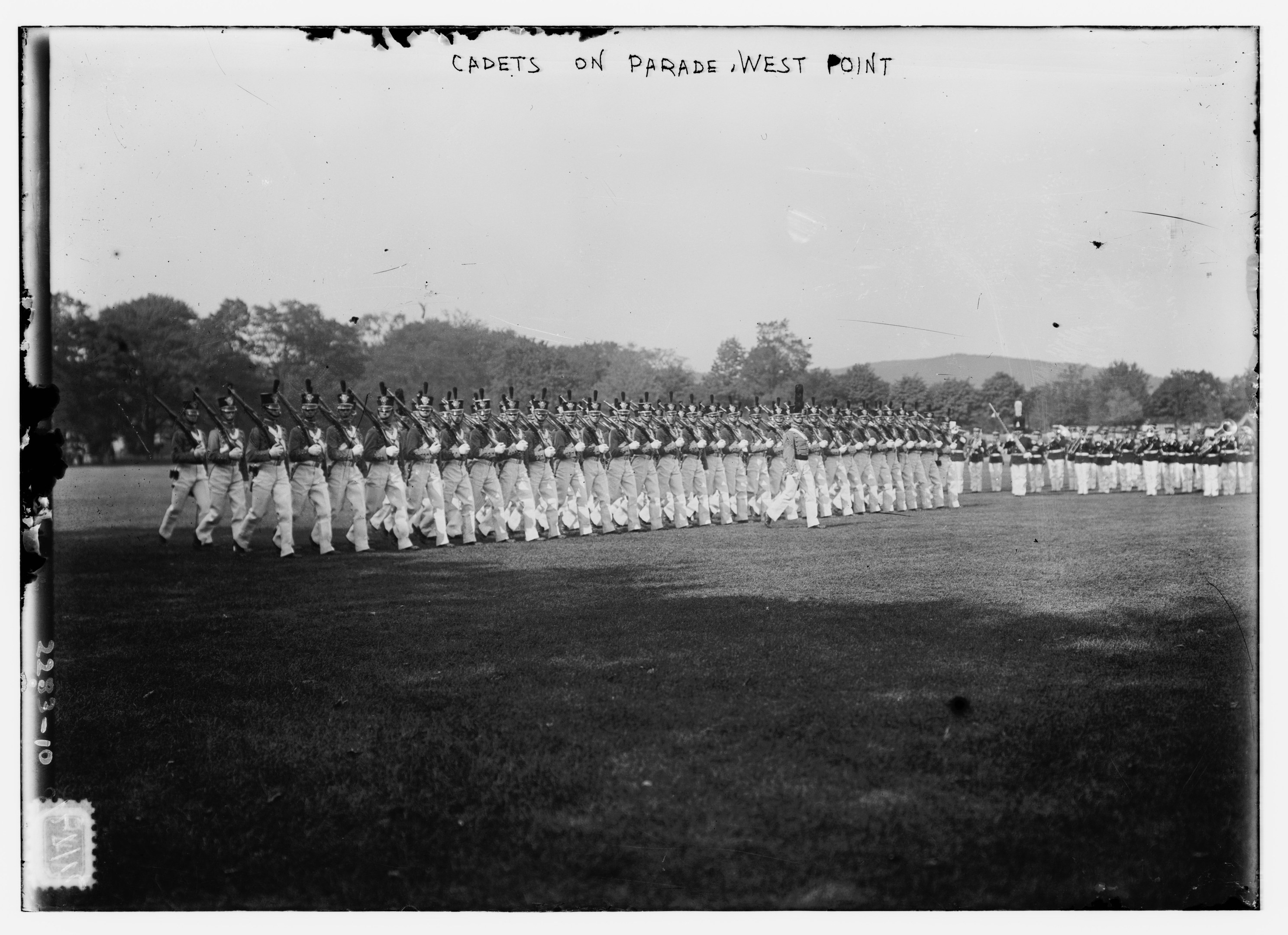 Gruppe von Menschen auf dem Boden stehend, einige halten Gewehre, mit Bäumen, Hügeln und einem bewölkten Himmel im Hintergrund; Text oben lautet "cadets on parade west point".