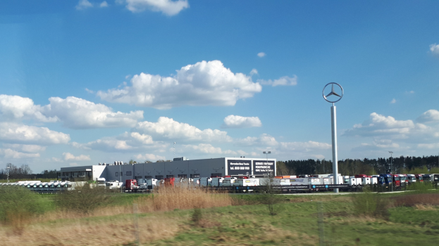 Eine Mercedes-Benz-Fabrik in Deutschland mit Fahrzeugen auf der Straße, Grünflächen, Gebäuden und einem bewölkten Himmel.