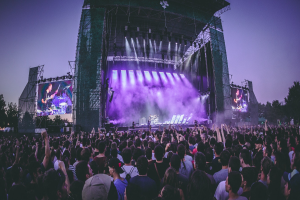 Eine große Menge Menschen bei einem Musikfestival schaut auf einer beleuchteten Bühne mit Instrumenten und Bildschirmen, die vor einem Gebäude, Bäumen und einem klaren blauen Himmel aufgebaut sind.