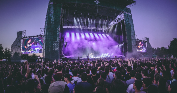 Eine große Menge Menschen bei einem Musikfestival schaut auf einer beleuchteten Bühne mit Instrumenten und Bildschirmen, die vor einem Gebäude, Bäumen und einem klaren blauen Himmel aufgebaut sind.