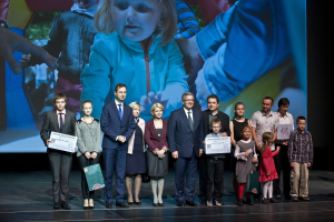 Eine Gruppe von Menschen auf einer Buhne mit Taschen und Urkunden, im Hintergrund ein Bildschirm mit einem Kind und anderen.