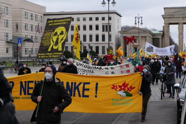Eine große Gruppe von Menschen marschiert auf einer Straße bei einer Demonstration gegen Atomkraft in Deutschland, mit Transparenten und Fahnen, mit Fahrzeugen auf der rechten Seite und Gebäuden, Laternenpfählen, Bäumen und einem Tor im Hintergrund.