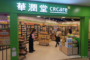 Eine Gruppe von Menschen steht vor einem Geschäft mit einer 'crcare'-Aufschrift, das Regale mit Lebensmitteln, Flaschen und Deckenlampen zeigt und eine Tafel trägt, die darauf hinweist, dass es Teil einer chinesischen Supermarktkette ist.