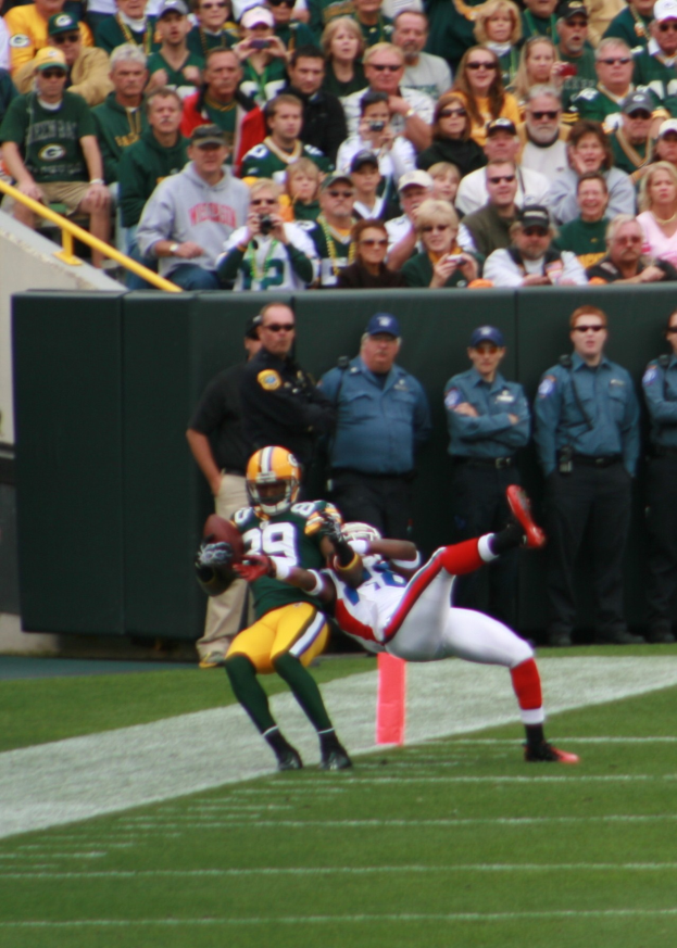 Ein F├╝├čballspieler f├Ąngt einen Ball vor einer Menge, die Sportbekleidung, Helme und Schuhe tr├Ągt, wobei einige Zuschauer Kameras halten und eine Wand im Hintergrund zu sehen ist.