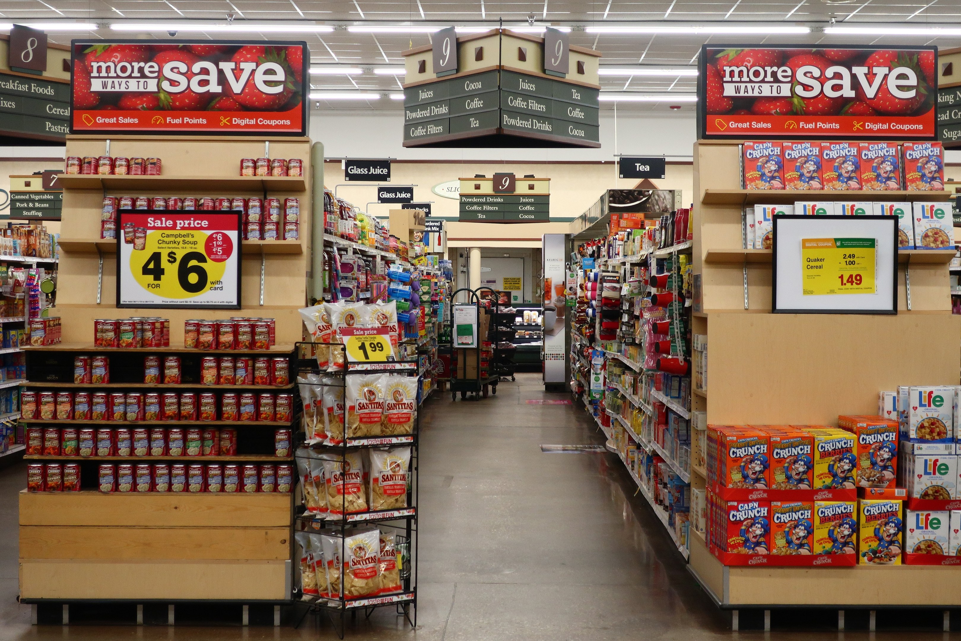 Ein Kiosk-Innenraum mit Regalen und Boden voller verschiedener Lebensmittel, Deckenleuchten und Informationsschildern, mit einer Person im Hintergrund.