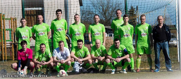 Eine Gruppe von Männern in grünen T-Shirts steht und hockt auf einem Fußballfeld mit einem Ball vor ihnen, ein Torpfosten rechts und ein Hintergrund aus Bäumen, Gebäuden, Polen, Fahrzeugen und dem Himmel.