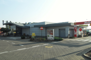 Ein McDonald's-Restaurant an einer Straßenecke, umgeben von Gebäuden, Schildern, Fahrzeugen, Infotafeln, Büschen, Bäumen und einem klaren blauen Himmel.