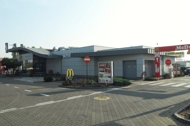 Ein McDonald's-Restaurant an einer Straßenecke, umgeben von Gebäuden, Schildern, Fahrzeugen, Infotafeln, Büschen, Bäumen und einem klaren blauen Himmel.