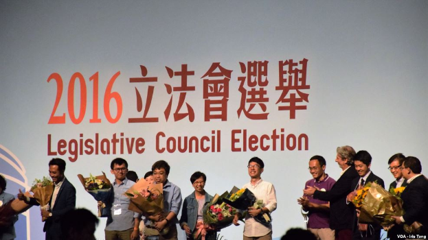Eine Gruppe von Menschen auf einer Bühne mit Blumensträußen, darunter ein Publikum und ein Bildschirm mit Text und Logo für die Wahl des Legislativrats 2016 im Hintergrund.