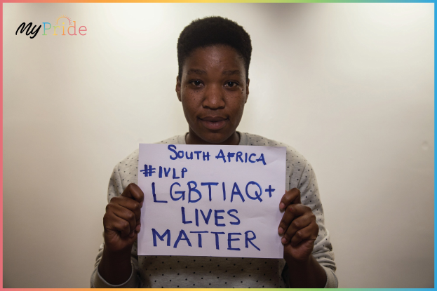 Eine Frau in einem weißen Hemd hält ein Schild mit der Aufschrift "LGBTQ+ Lives Matter" mit entschlossener Miene vor einer Wand.