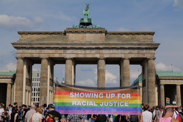 Eine Gruppe von Menschen mit einer "Racial Justice"-Schlagzeile vor dem Brandenburger Tor in Berlin, Deutschland, mit Gebäuden und einem bewölkten Himmel im Hintergrund.