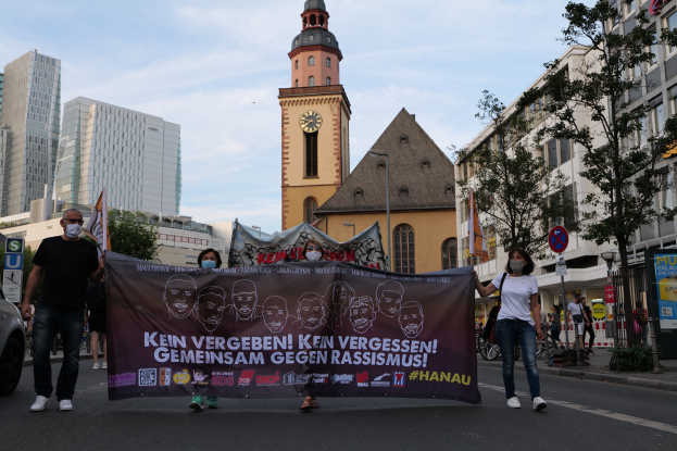 Eine Gruppe von Menschen in Masken, die eine Straße entlanggehen und ein Banner mit Text und Bildern halten, mit einem geparkten Auto auf der linken Seite, Gebäuden und Bäumen im Hintergrund und einem Uhrenturm unter einem klaren blauen Himmel.