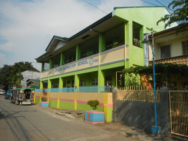 Ein Schulgebäude mit grüner und gelber Fassade neben einer Straße, mit Fahrzeugen, Topfpflanzen, einem Zaun, einem Tor, einem Straßenpfahl, Bäumen, Drähten und einem bewölkten Himmel.