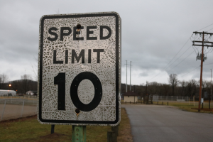 Geschwindigkeitsbegrenzungstafel am Rande einer Straße mit umliegender Wiese, Strommästen, einem Zaun, Häusern, Bäumen und einem bewölkten Himmel im Hintergrund.