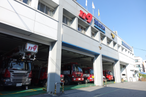 Eine Feuerwache mit mehreren geparkten Feuerwehrautos, mit Fenstern, Säulen und Namensschildern, mit Flaggen im Vordergrund und anderen Gebäuden unter einem klaren blauen Himmel im Hintergrund.