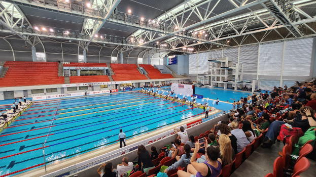 Großer Hallenbad mit vielen Menschen auf Stühlen sitzend und einigen im Wasser stehend, mit hoher Decke mit Lampen und Eisenstangen und Schildern mit Text im Hintergrund.