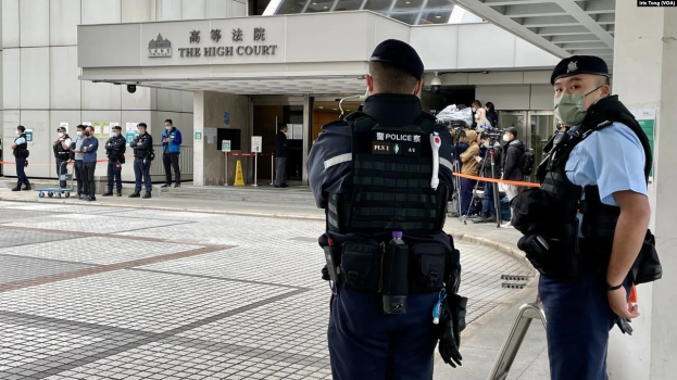 Zwei Polizisten in Uniform, Mützen und Masken vor einem Gebäude am Hong Kong International Airport, umgeben von Menschen, einige mit Kameras, mit Säulen im Hintergrund, die mit Bändern geschmückt sind.