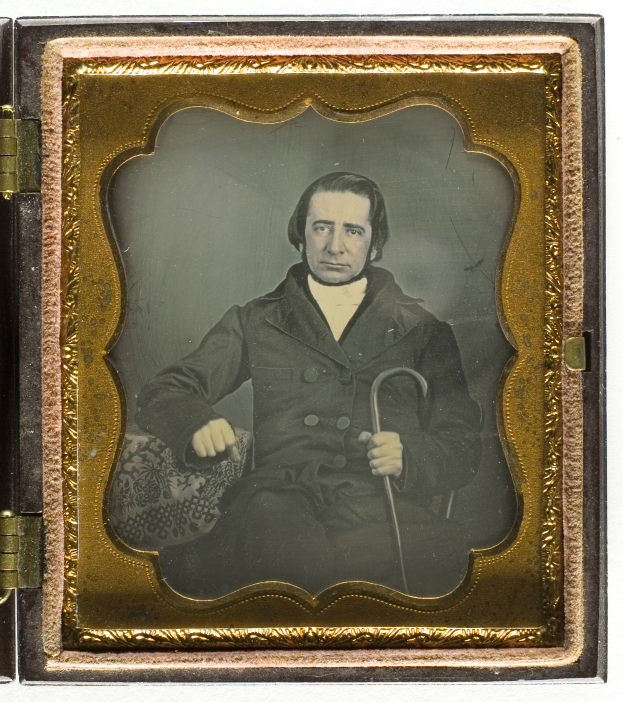 Ein altes Daguerreotyp eines Mannes in einem Anzug, der einen Stock hält und ernst in die Ferne schaut.