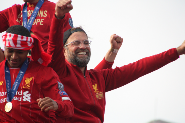 Liverpool-Trainer Jürgen Klopp feiert mit seinen Spielern den UEFA-Champions-League-Sieg, während zwei medalletragende Personen in der Nähe sind und der Himmel im Hintergrund zu sehen ist.