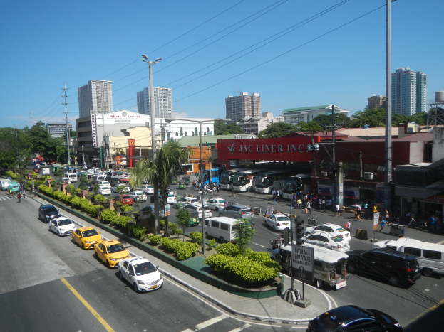 Eine belebte Stadtstraße mit zahlreichen Autos, Bussen und Fußgängern, umgeben von Gebäuden, Bäumen und Straßeninfrastruktur unter einem klaren blauen Himmel.