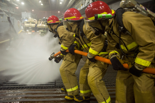 Feuerwehrleute in Helmen und Handschuhen halten Schläuche und spritzen Wasser auf ein Löschfahrzeug, mit Lichtern, Geländern und Tafeln mit Text im Hintergrund.