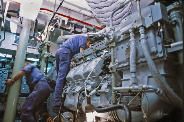 Zwei Männer in blauen Uniformen arbeiten an einem großen Motor in einer Fabrik, umgeben von verschiedenen Maschinen und Geräten.